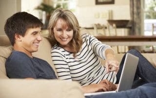 mother with teenage son sitting on sofa at home using laptop