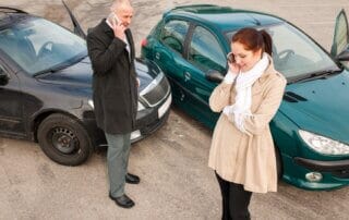 alt="A man and woman on their phones standing along side their black and green car that collided.".
