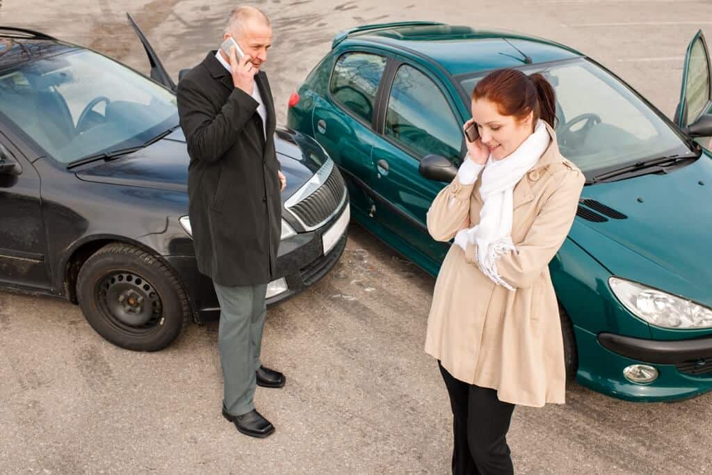 alt="A man and woman on their phones standing along side their black and green car that collided.".