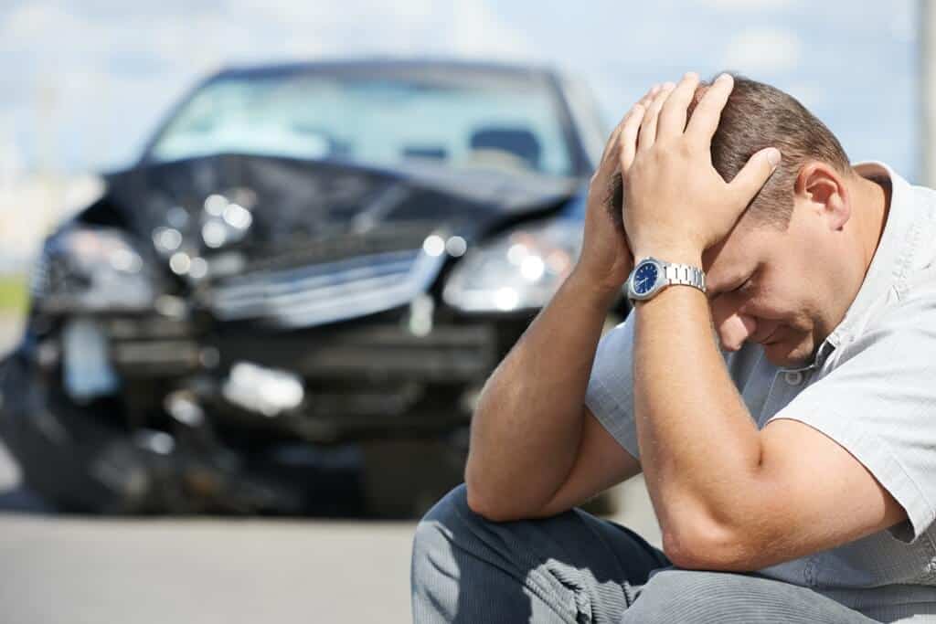 alt="Man sitting down by the road with both hands on the head with his black crushed car on the side."