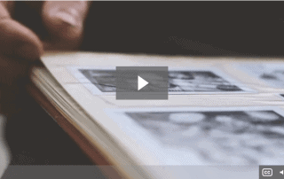 alt="Old black and white photos in the album with a hand trying to flip the page and a play video sign in the center."