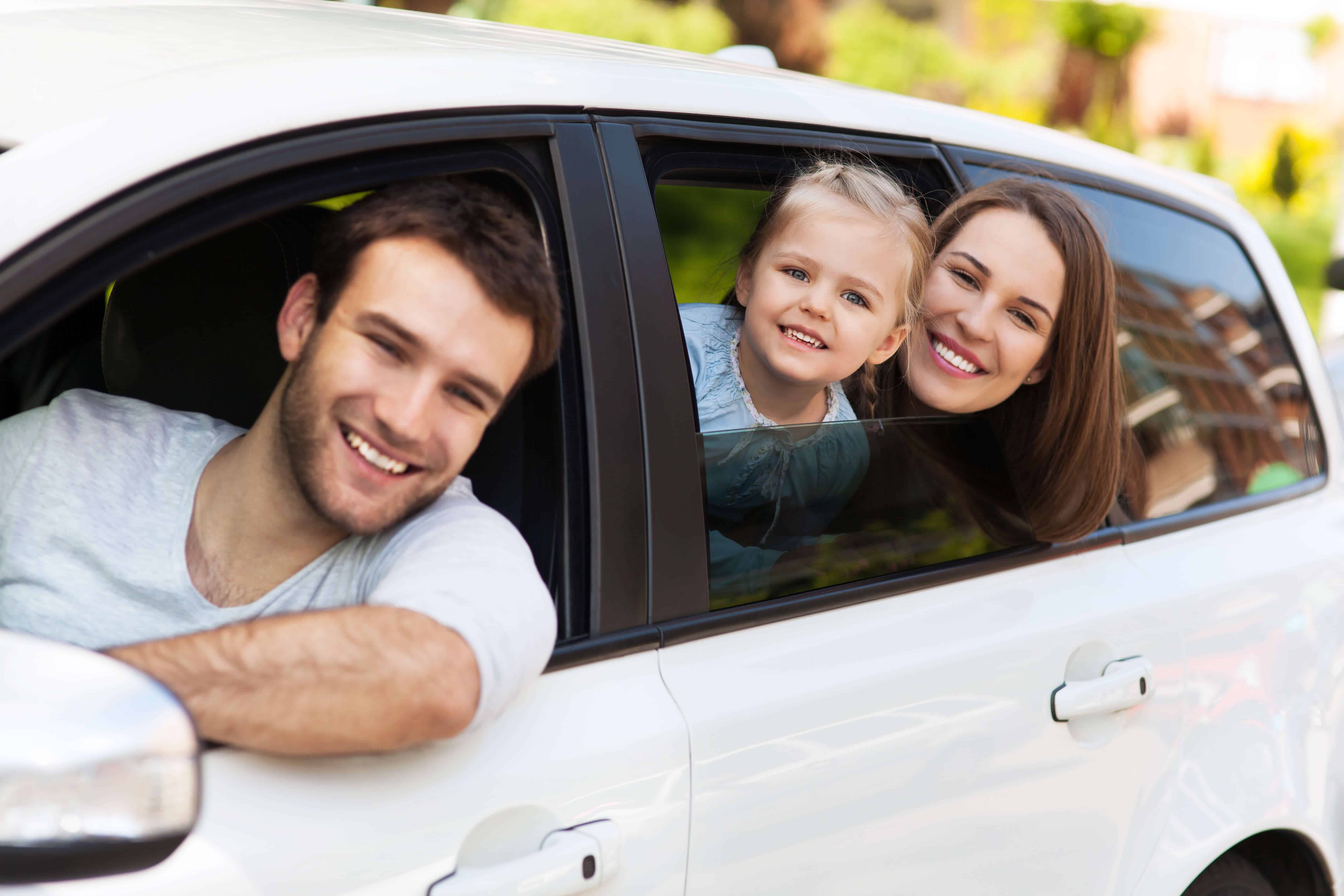 alt="White car with a family, the father is the driver, they're all looking out the window smiling."