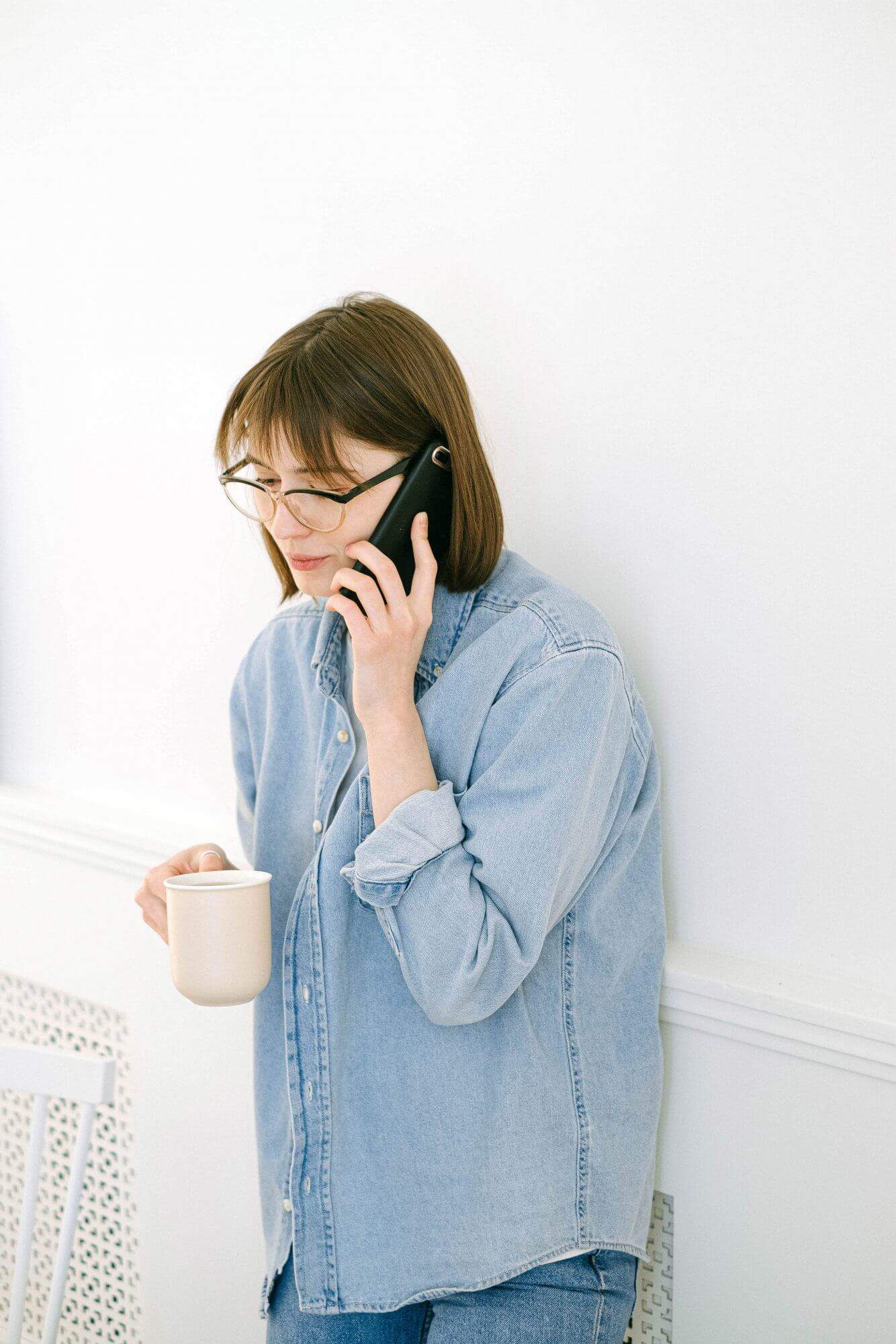 alt="A woman in denim shirt and pants talking to an insurance advisor over the phone, holding a white coffee mug."